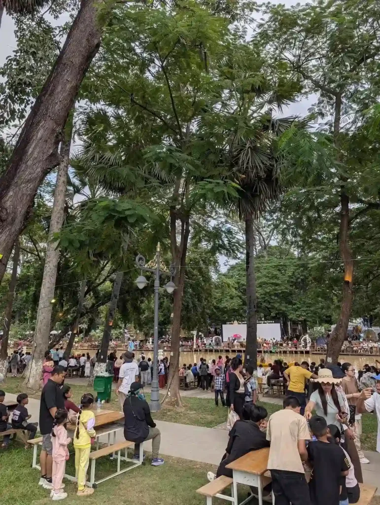 An image of a crowd, taken further back along the the shore of the Siem Reap river during the Boat Racing festival (Bon Om Touk) in Siem Reap, Cambodia. There are lots of trees. People are sitting at picnic tables near the sidewalk, others are standing around, and others are sitting in the grass next to the river. Most people are watching the boats racing, but others are eating food and socializing. In this image, the river can be seen in the near background, but there are no boats racing in this image.