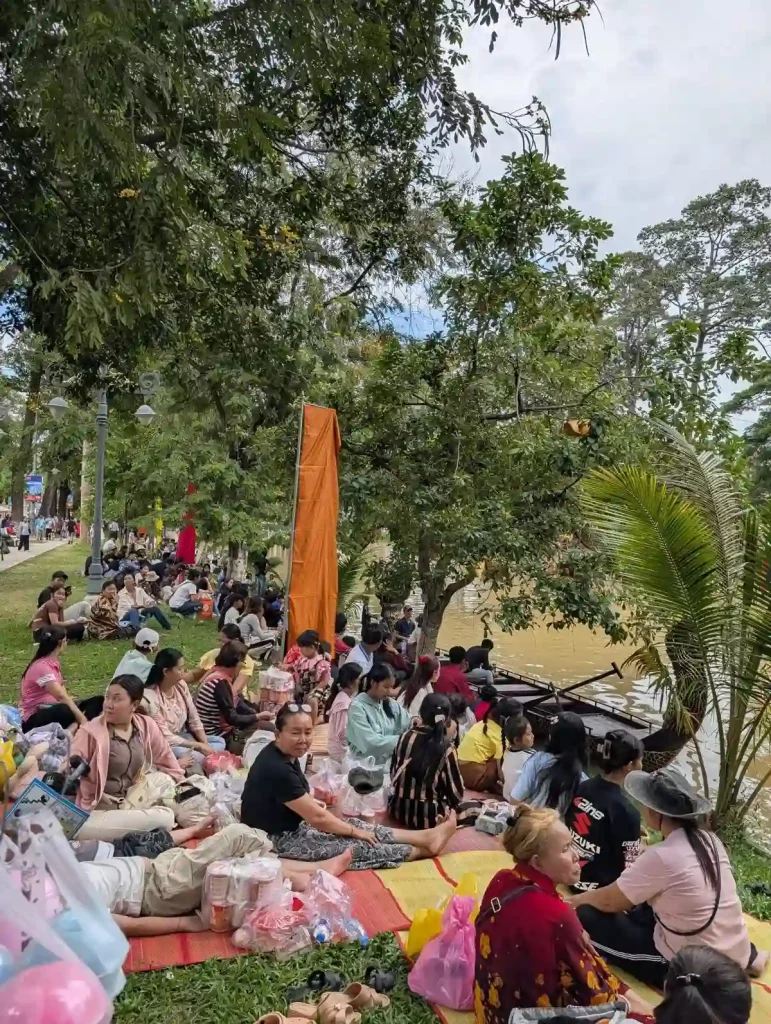 An image of a crowd along the the shore of the Siem Reap river during the Boat Racing festival (Bon Om Touk) in Siem Reap, Cambodia. People are sitting in the grass on mats socializing and watching the boats racing. In this image, the river can barely be seen, and neither can the boats racing.