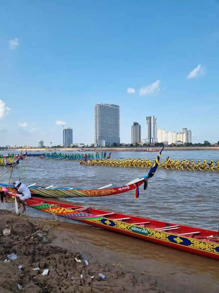 An image of the Boat Racing festival (Bon Om Touk) in Phnom Penh, Cambodia. This is taken on the shore of the Mekong river next to a couple of parked boats. There is a race between two boats in the river in the background. In the far background are some high rise buildings. The Mekong river is very wide at this point.