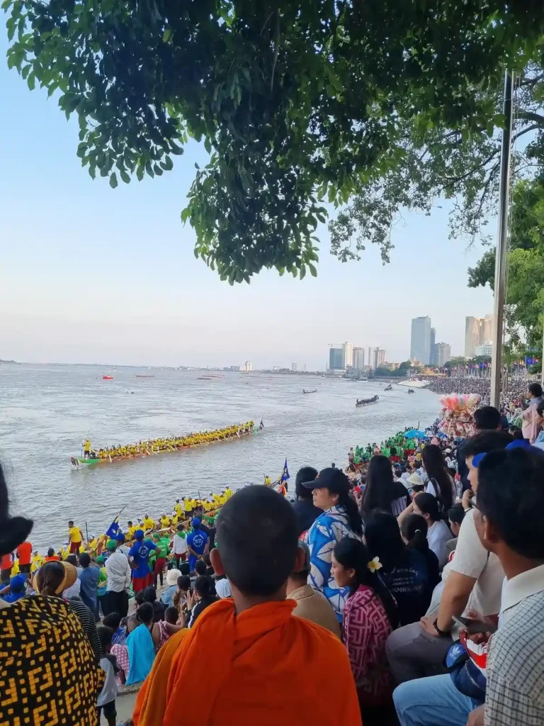 An image of the Boat Racing festival (Bon Om Touk) in Phnom Penh, Cambodia. This is taken at an elevated point on the shore of the Mekong river, with a packed crowd, watching the long boats racing in the river below. The boats are paddled by roughly fifty men. In the foreground is a Buddhist monk in Kasaya (a monk's robe colored in saffron dye). The Mekong river is very wide at this point.