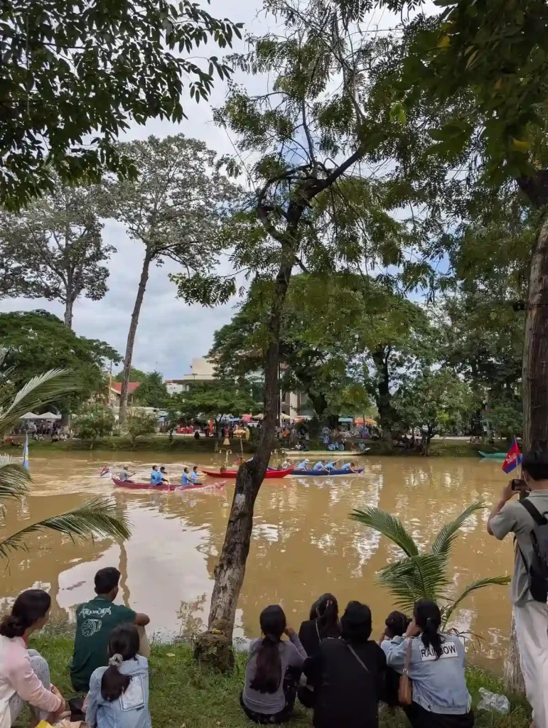 An image of the Boat Racing festival (Bon Om Touk) in Siem Reap, Cambodia. This is taken on the shore of the Siem Reap river, with people sitting in the grass watching the boats racing in the river. The boats are paddled by four to six men, and two boats are racing.