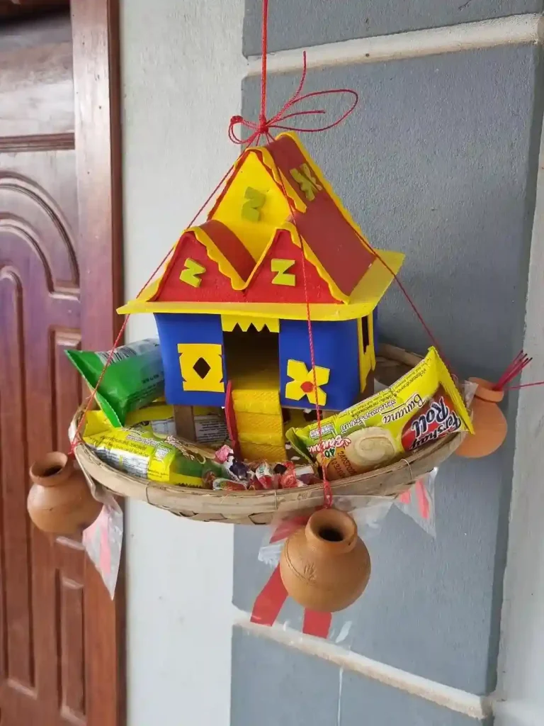 A small child-version spirit house hanging next to a front door of someone's home. This spirit house has snacks (sweet roll desert and a few candies), which is intended for the child spirits.
