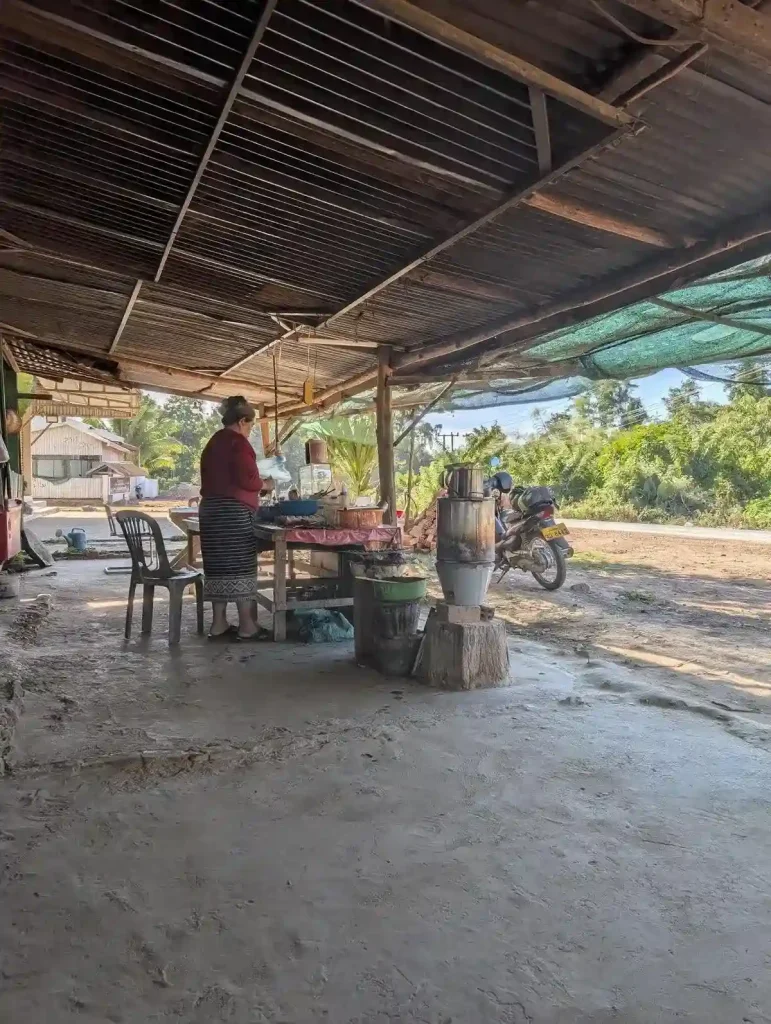 A Laos woman tending a roadside shop selling coffee and snacks in a small village in Laos. The shop has a concrete floor that is not smooth, and a corrugated metal roof held up by wooden framing. This is more like the patio of someone's house that they decided to use as a shop. A motorbike is parked in front of the shop.
