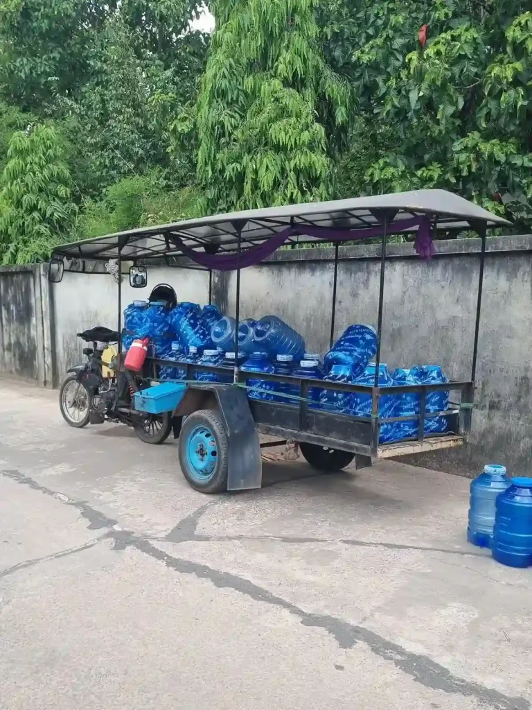 A delivery man with a load of twenty liter water jugs of filtered water in Southeast Asia. The load of water jugs are loaded in a trailer with a roof covering, and towed with a motorbike.