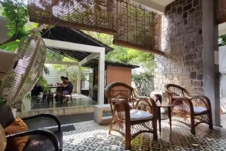 Areas to hangout at a guesthouse in Southeast Asia. The area is open-air with a roof covering. There are chairs and tables with some outdoor space for relaxing.