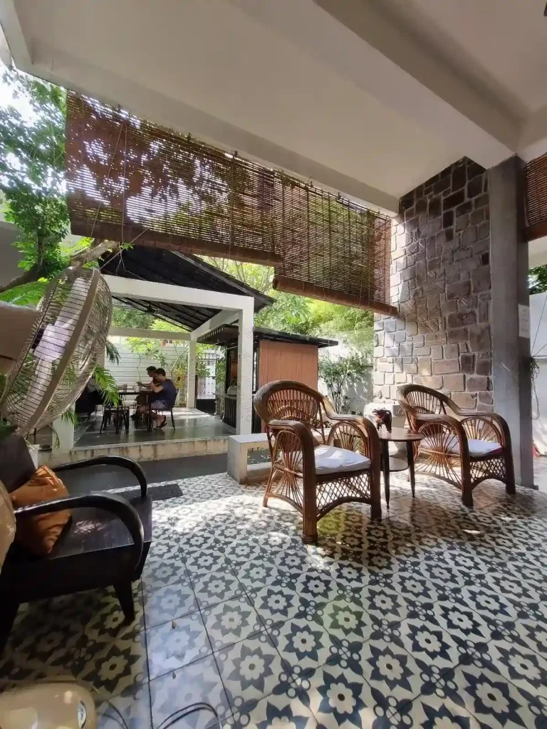 Areas to hangout at a guesthouse in Southeast Asia. The area is open-air with a roof covering. There are chairs and tables with some outdoor space for relaxing.