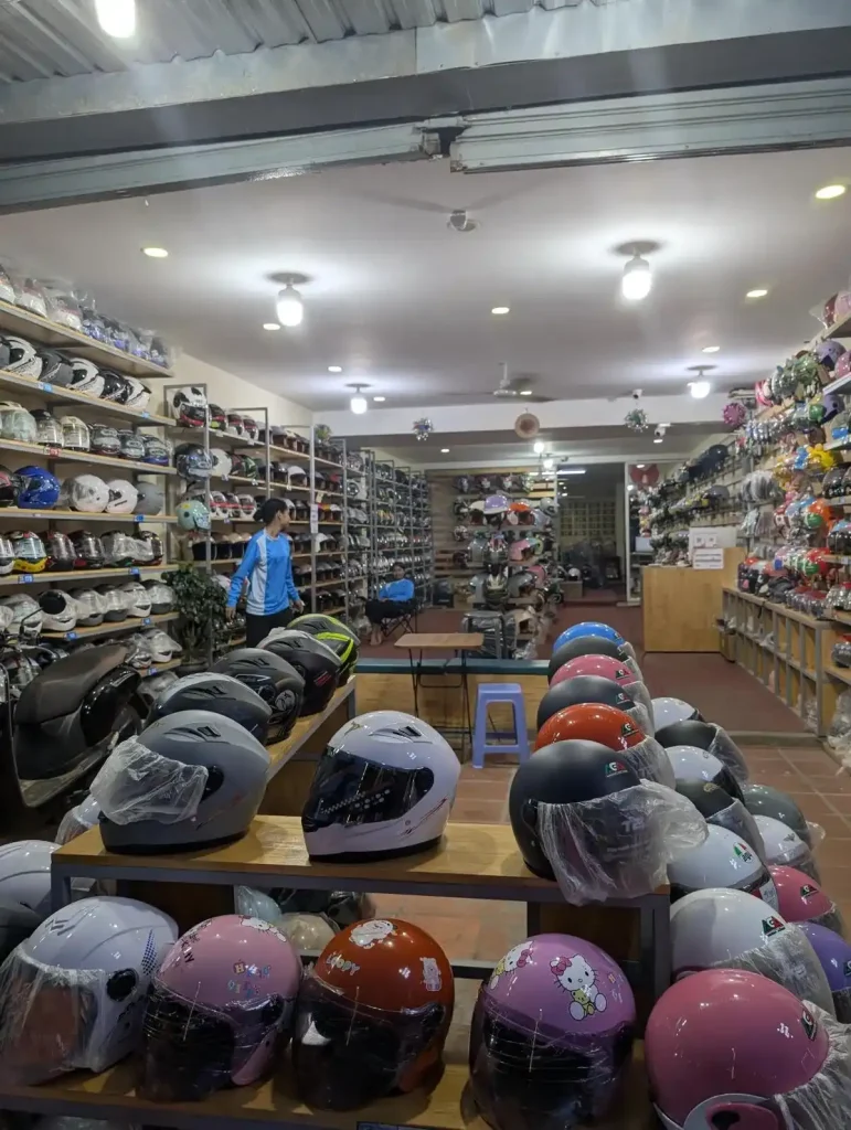A storefront with motorcycle helmets in Southeast Asia. In the foreground is a display of helmets on display at the entrance of the store. In the background, the walls are covered with shelving that are fully stocked with helmets on them. The helmets are of various styles. The helmets in the image foreground have a face shield feature that can be pulled downward (to use the face shield) or upward (to get rid of the shield). Some have chin coverage, others do not (i.e. not a full-face-covered helmet).