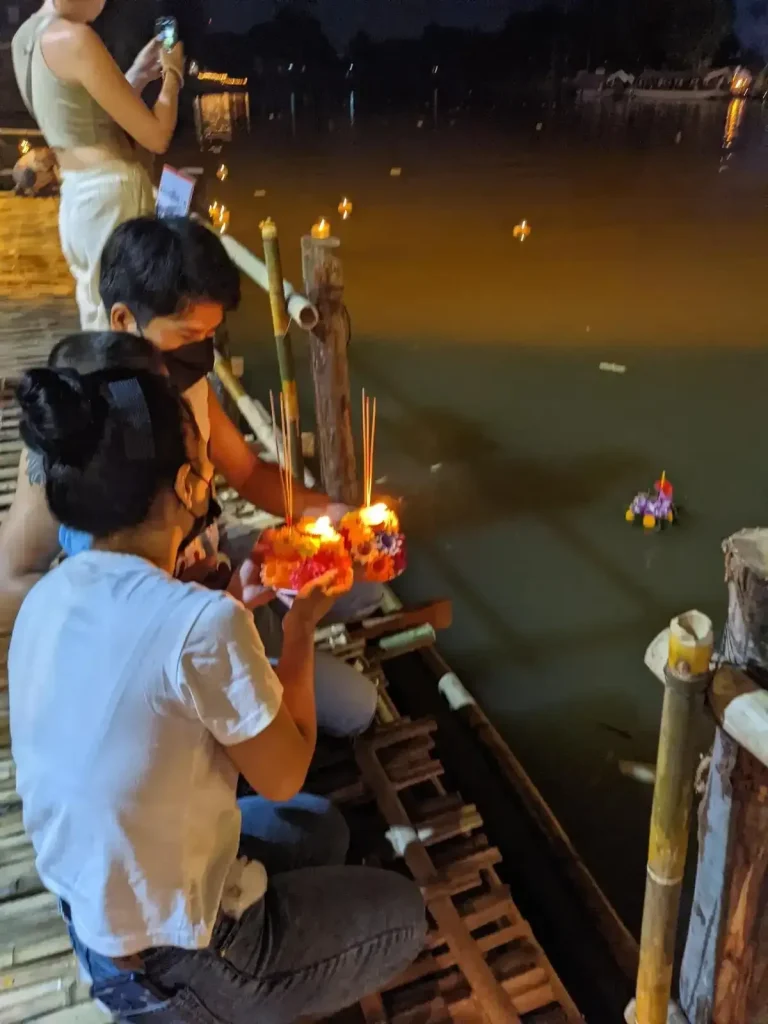 A male and female each holding a krathong and placing them into the river during Loi Krathong holiday in northern Thailand. This is at night, and the krathongs have a lit candle in them, which glows while they float in the water. Several krathongs can be seen in the background in the water.