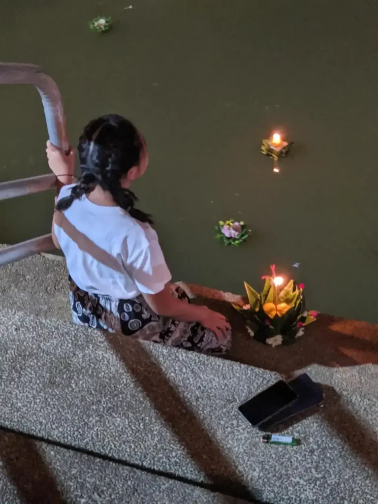 A young child with a krathong sitting next to them at the end of stairs that lead into the river water. They are about to place the krathong into the river as part of celebrating the Loi Krathong holiday in northern Thailand. This is at night, and the krathong has a lit candle within it, which glows while it floats in the water. A few krathongs can be seen that are already placed in the water.