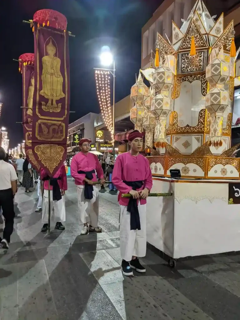 Men in a traditional Thai clothing, walking in a parade with a large lantern that celebrates the Lantern festival (Yi Peng holiday) in northern Thailand. This lantern is large, is on a small float with wheels, and four men are required to push it down the street. There are other men holding banisters with what appears to be a Buddha image on it and some decorative pattern.