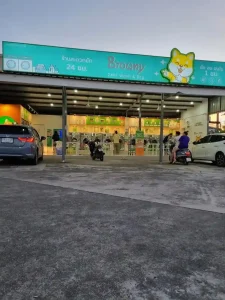 A laundromat building in Southeast Asia. It is located with a parking lot, and has two cars and two motorbikes parked under an awning roof covering. The laundromat is open-air on two sides. The other two sides have wall where the machines are mounted. There is a roof covering the building and entrance.