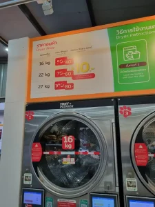 A clothes drying machine at a laundromat in Southeast Asia. The drier in the image is a front-loader machine, and is rated for a sixteen kilograms of clothing. There is a sign above the drier with prices: sixteen kilogram drier is fourth Thai Baht (about one dollar and twenty-five US cents), twenty-two kilogram drier is sixty Thai Baht (about two US dollars), and a twenty-seven kilogram drier is eighty Thai Baht (about two dollars and fifty US cents).