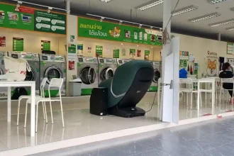 A laundromat with washing and drying machines in Southeast Asia. There are a few desks with chairs. Also, there is a well-padded massage chair for use. People take off their shoes when entering. The laundromat is open-air on two sides. The other two sides have wall where the machines are mounted. There is a roof covering the building and entrance.