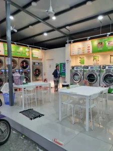 A laundromat with washing and drying machines in Southeast Asia. There are two desks that each have four chairs. People take off their shoes when entering. The laundromat is open-air on two sides. The other two sides have wall where the machines are mounted. There is a roof covering the building and entrance.