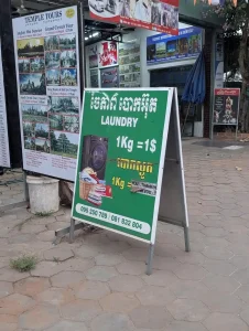 A sign for a laundry service on a sidewalk in Southeast Asia. This service will wash and dry your clothes, bed sheets, and more. Typically drying is hang dried, but some services have drying machines.