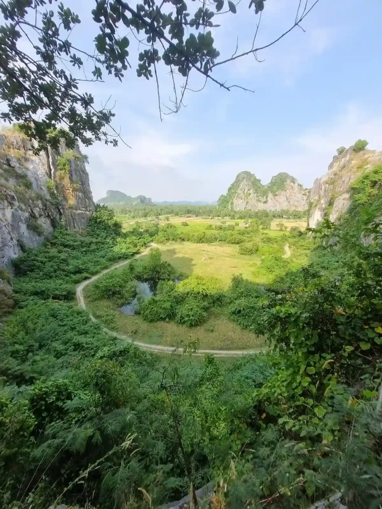 A viewpoint seen after hiking to the top of a mountain in Southeast Asia. The image was taken at a cliff. Below is a valley surrounded by the mountain. Below, on the ground is a dirt road that goes in a circle around the valley.