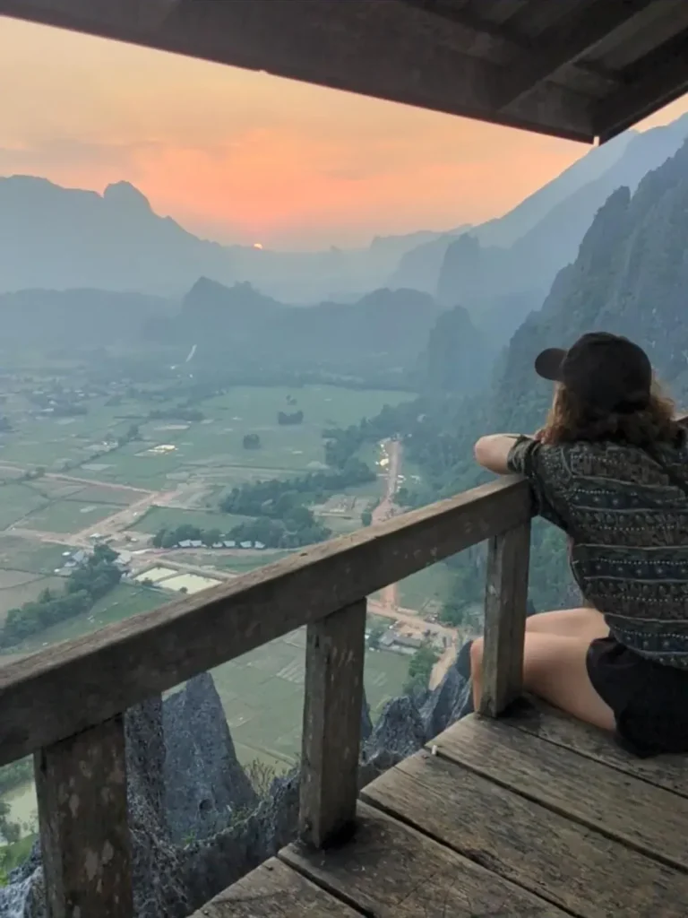 A mountain hiking viewpoint with sunset at Nam Xay Viewpoint in Vang Vien, Laos. The image is within a roof-covered wooden observational deck at the top of a mountain peak. A female is at the edge of the deck, with her legs dangling off the edge, and arms resting on the short railing. Her chin is resting on her arms. She is peacefully soaking in the view and sunset. It is quite at the top.