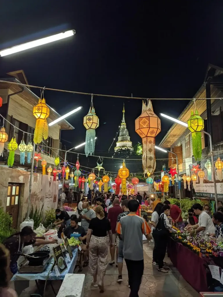 A temporary outdoor night market along a walkway during a holiday in Thailand. There are lanterns hanging above the walkway, and the market is busy with people. For sale are some snacks, handicrafts, and kratongs (a flower origami-type thing that is put into the water with a candle during the Loi Krathong holiday. The holiday celebrated is actually two holidays at the same time, Loi Krathong (involves hand made krathongs) and Yi Peng (lantern festival).