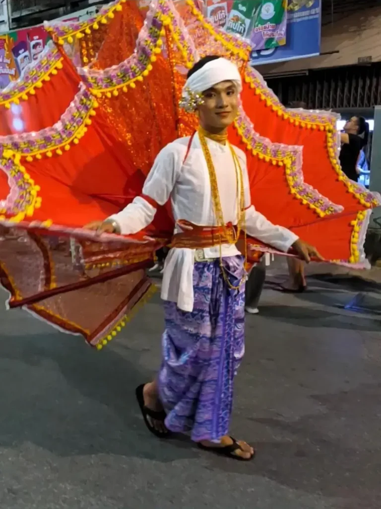 A male in a traditional Thai costume, walking in a parade that celebrates the Lantern festival (Yi Peng holiday) in northern Thailand. A prop that is similar to a turkey tail is mounted to his waist. It is controlled with his arms like wings of a bird, and spans over his head and double his arm length. It is decorated. This turkey tail is described as being large and hemisphere-shaped that resembles a hand-held chinese fan.