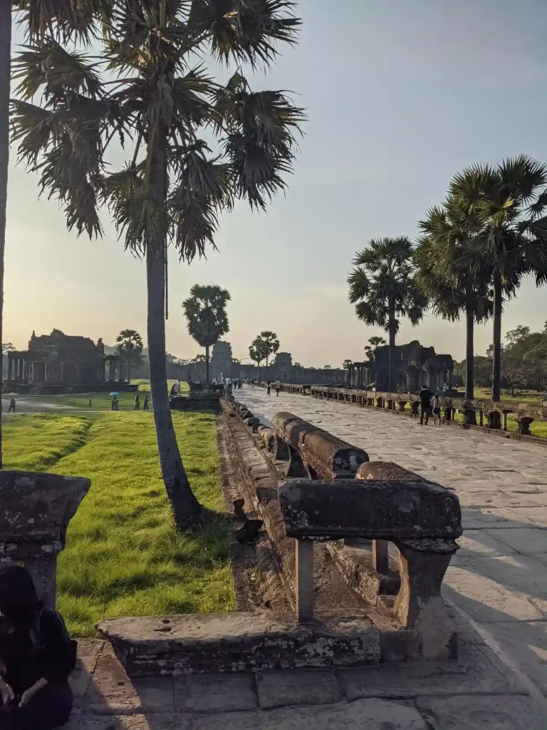 The road leading to and within the Angkor Wat temple complex grounds. This is located within Angkor Archaeological Park, which is a UNESCO World Heritage Site. This image does not show Angkor Wat, but from the road looks towards the west entrance gate of the complex. There is a small building to the left and right, in which this road goes between. A palm tree is in the foreground next to the road.