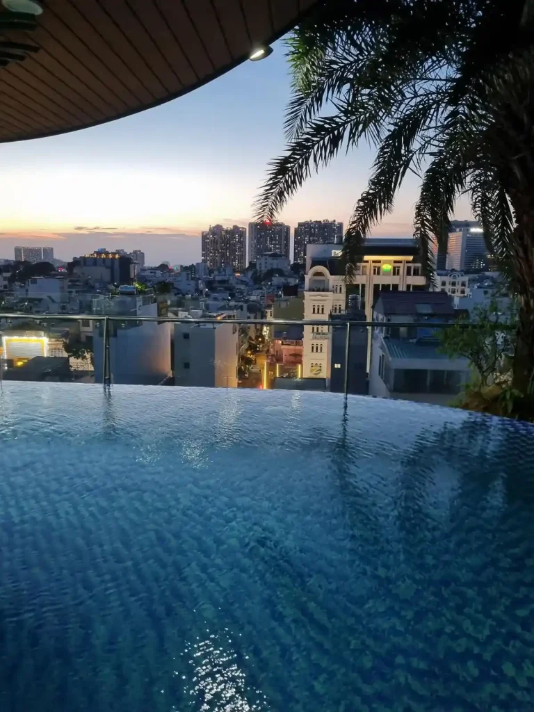 Viewing a sunset from a rooftop infinity swimming pool in Southeast Asia. High rises can be seen between the pool and the sunset, since this takes place in a large, densely populated, city in Southeast Asia.
