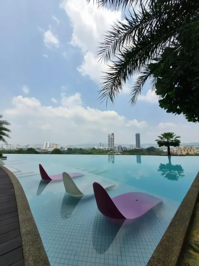 Three chairs inside a apartment rooftop swimming pool in Southeast Asia. The pool faces a view with no buildings in it's way. In the background are a few high rise buildings and tree tops of trees below.