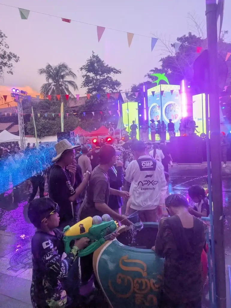 A local concert during the Songkran holiday festival in Cambodia. Two local boys are refilling their waterguns at a water basin in the foreground - one is shooting his as someone off the image. Because a continual water fight and splashing of water is part of this festival, there is water being sprayed everywhere in the crowd.
