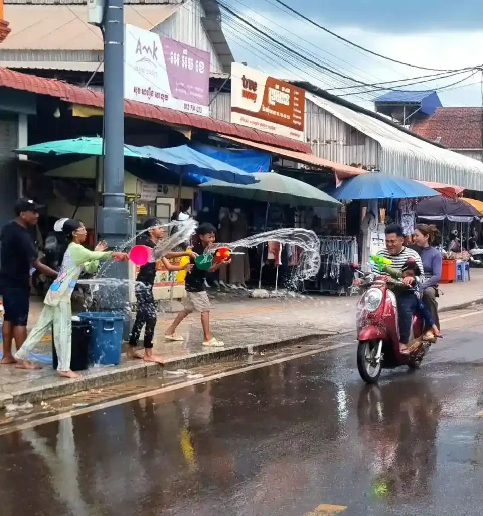 Three kids splashing a motorist (husband, wife, and child) on a scooter with water during the Songkran holiday festival in Cambodia. Everyone is smiling. The motorist is riding by on the street, and the kids use pales of water to spash them. They have a large bucket of water to refill their pales with. This festival is essentially one massive water fight, and everyone gets soaking wet.