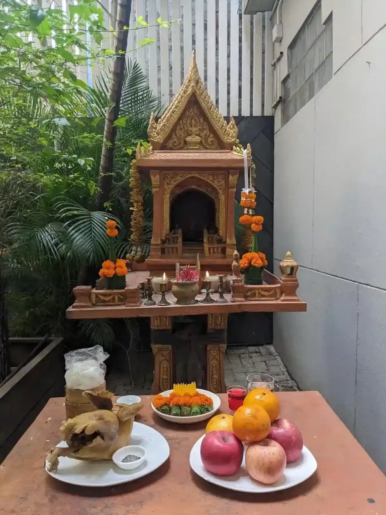 A spirit house with several offerings including a whole boiled chicken, three apples, three oranges, a large portion of sticky rice, some deserts, cup of water, a shot of red alcohol, a few flowers, burned incense, and two small candles.