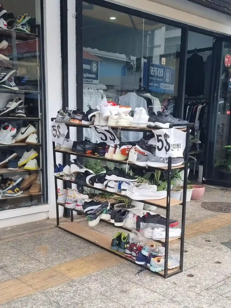A store selling used shoes in Cambodia. This image shows a rack of used shoes, including several pairs of name brand Nike, Adidas, and New Ballance shoes in good condition. There is a sign on the rack that references the price of five US dollars. FYI: the US dollar is an official currency in Cambodia, along with the Khmer Riel. So, in Cambodia, prices are sometimes listed in US dollars.
