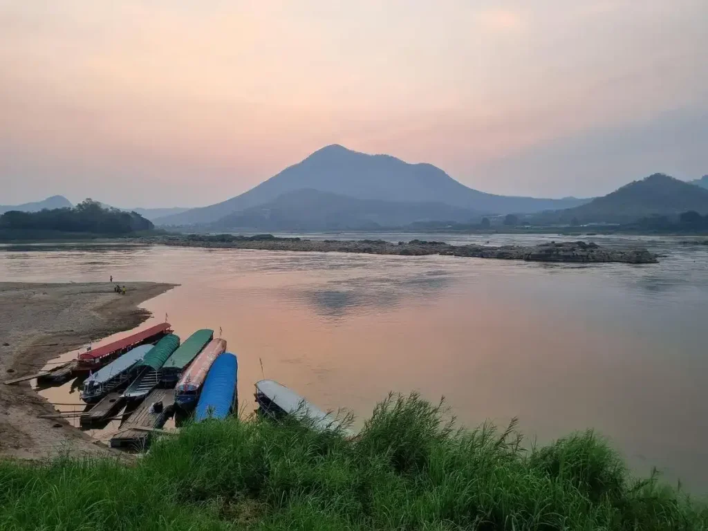 A sunrise on the Mekong river, which serves as the border line between Thailand and Laos. The sun is still behind the mountains beyond the river, but light can be seen. There are seven long boats docked along the shore, together. This image was taken on the Thailand side facing Laos.