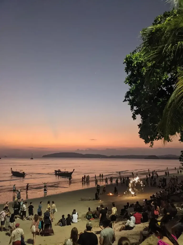 A sunset view at Ao Nang beach in southern Thailand. There are many people on the beach, but it is not overly crowded. There are two Thai longtail boats in the water near the shore. And, in the distance are low mountains (not too tall) where the land extends far into the ocean. The sunset can be seen with the mountains as a backdrop.