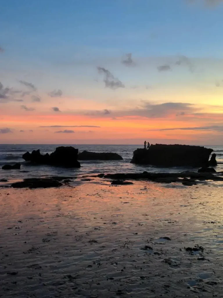 A sunset at a black sand beach in Bali, Indonesia. The sand is wet, and reflects the color of the sunset. In the middle of the image are one story high rock formations just off the near shore of the beach. Two people are standing one of the rocks formations. In the background is an organge sunset. From bottom to top, the orange sunset fades to yellow and then blue (at the top of the image).