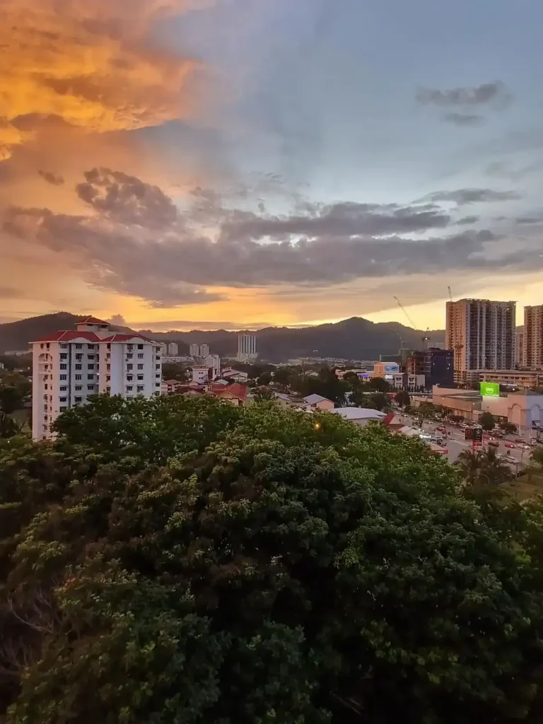 A sunset with mountains in the background. High rise housing is between the background and foreground, which are just slightly shorted than the mountains. In the foreground is the top of a tree, which takes up the entire width of the image. The sky is partially bright orange color and light blue. One cloud is light-up bright orange.