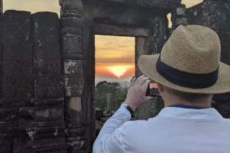 A man taking a photograph of a sunset within Angkor Archeological Park in Cambodia. There is an ancient doorway and wall made of stone between the man and the sunset. However, the photographer is capturing the sunset (can be seen in this image) through the doorway. The doorway has no door, so it is open.