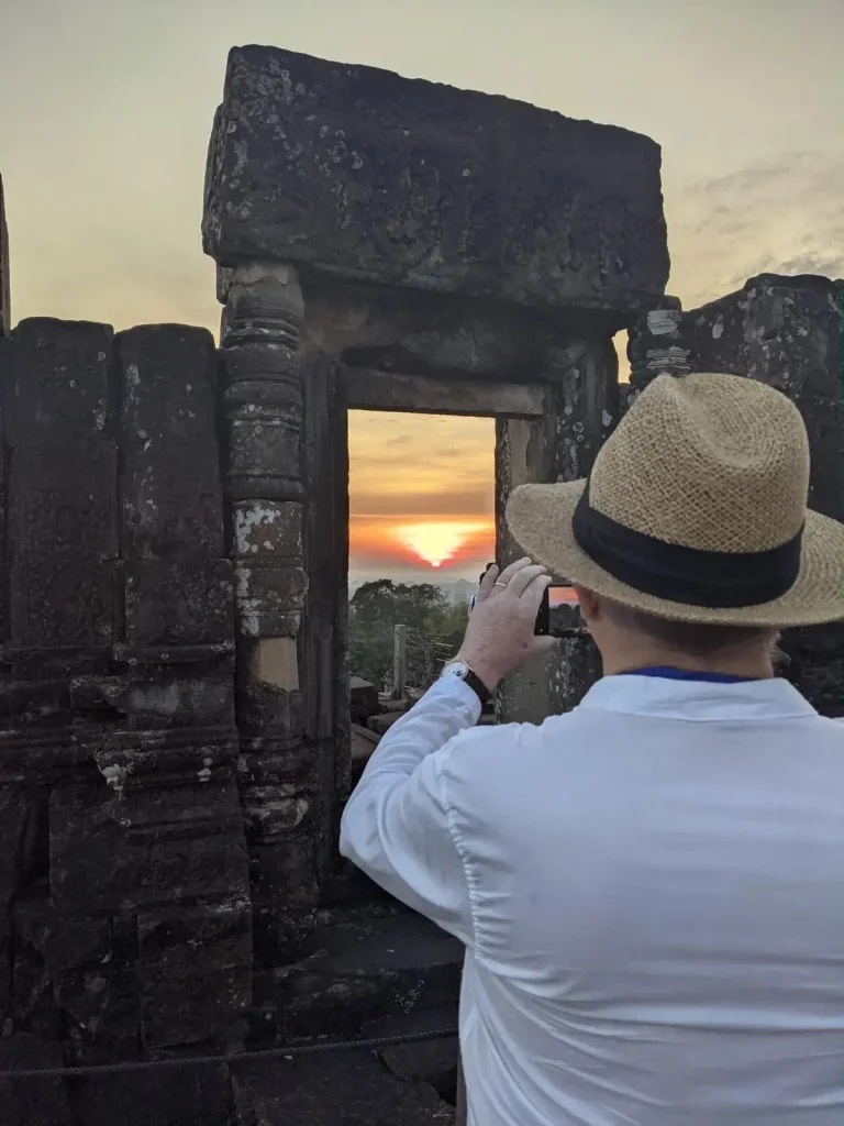 A man taking a photograph of a sunset within Angkor Archeological Park in Cambodia. There is an ancient doorway and wall made of stone between the man and the sunset. However, the photographer is capturing the sunset (can be seen in this image) through the doorway. The doorway has no door, so it is open.