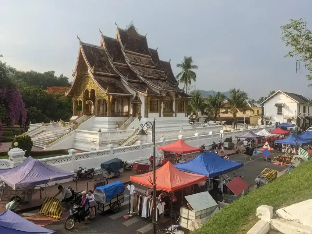 A night market being setup on a street outside a temple in Luang Prabang, Laos. This image is taken next to the market, looking down on it from the top of some steps. It shows the street with pop-up canopies being setup, and the temple on the other side of the road.