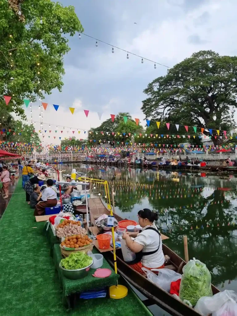 A festival in Thailand that is full of food booths. This image shows the part of the festival setup along a small, still, river. There is a food booth in the foreground, in which the seller is in a small boat (it is mounted to the sidewalk, but overhangs into the river). There are festival flags sprawling across the entire length of the river. These flags are triangle shaped, of different solid colors, and attached to a rope that is connected to elevated points on both sides of the river.