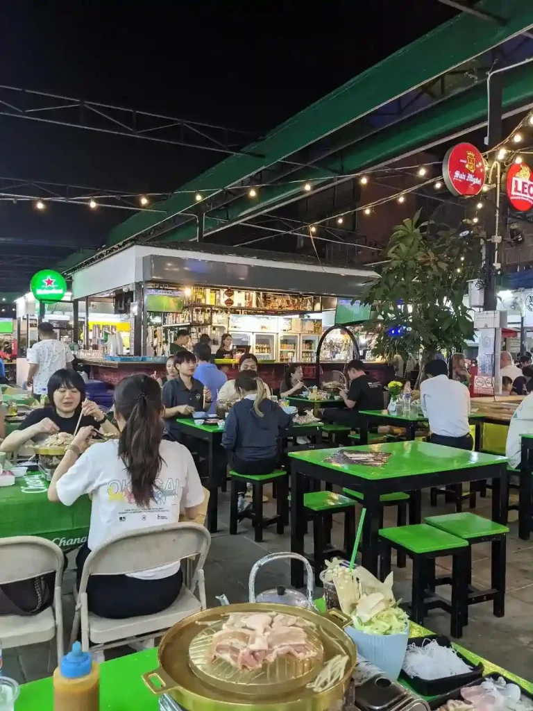 A typical outdoor Thai-style market, specifically for having a sit down meal at. There are tables & chairs and people eating. This is similar to a hawker area in Malaysia & Singapore. Barely noticeable in this image, is how there are multiple vendors, which are essentially different restaurants.
