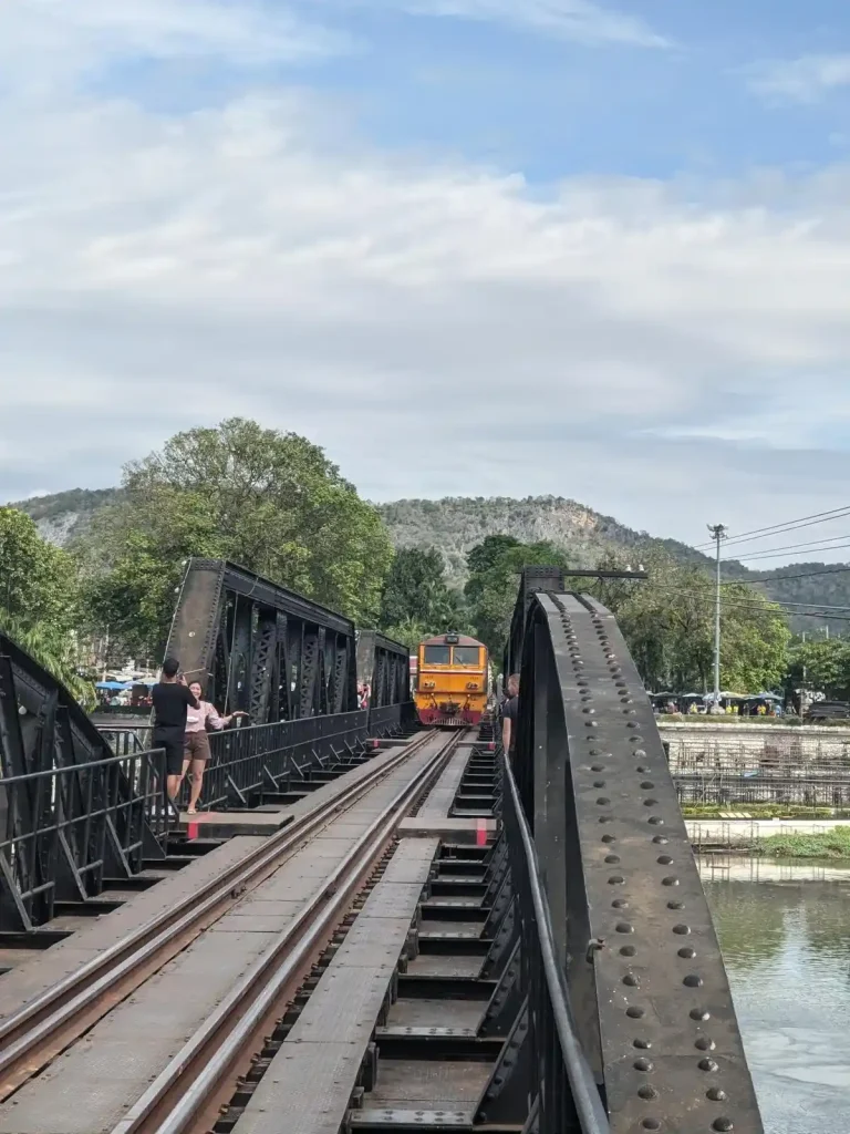 On the River Kwai Bridge in which the tourist train runs on. There is a train on the bridge, slowly approaching the photographer, who is on the tracks. However, there are viewing decks on the bridge for people to wait on as the train slowly passes by. This has world war two significance, and is located in Kanchanaburi, Thailand.