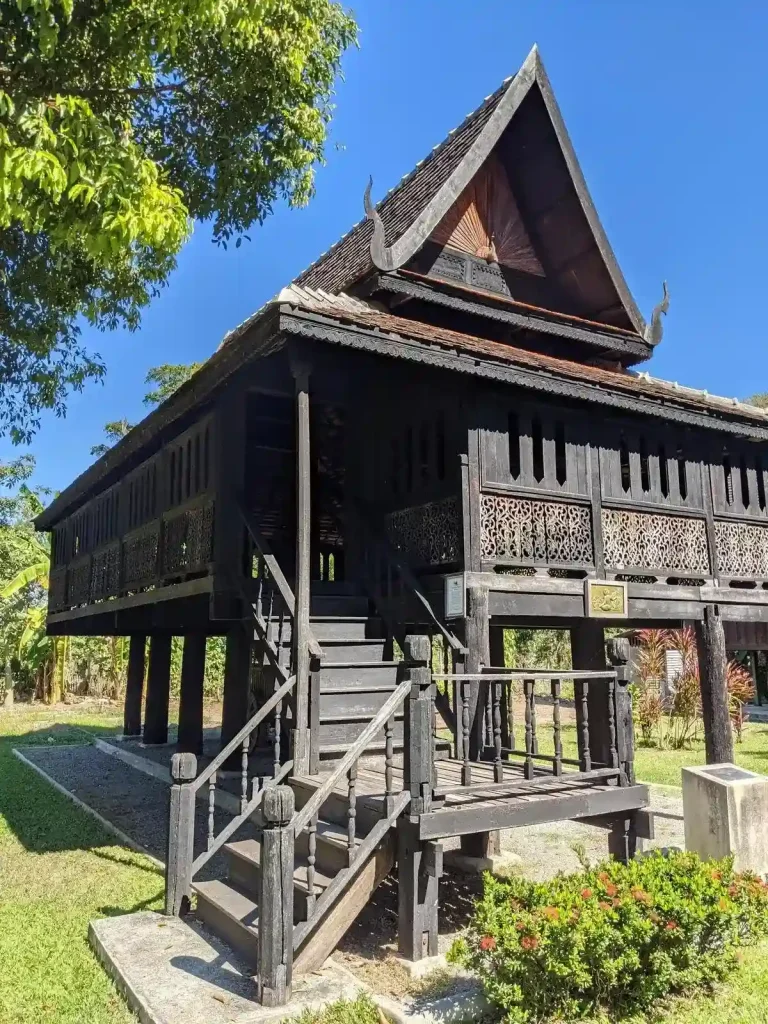 A traditional northern wooden stilt house in Thailand. The home has vertical wooden beam mounted in the ground, and an elevated platform (the floor of the home) attached to it. There are stairs leading up to the home from the ground below. This is part of a museum in Chiang Mai, Thailand.
