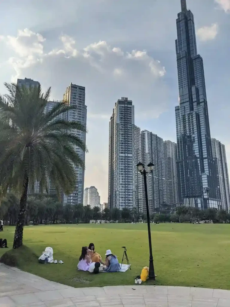 An image taken in a park at a Vin Home complex in Saigon (aka Ho Chi Minh City), Vietnam. The park is surrounded by condo high rises. There is a group of three females sitting on a mat in the park. Nearby them is a palm tree. The park is well maintained.